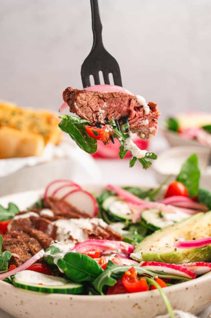 A black fork lifting a bite of steak with salad out of a bowl.