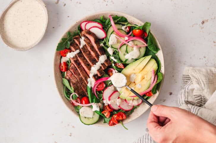 A hand spooning creamy dressing over a salad with steak.