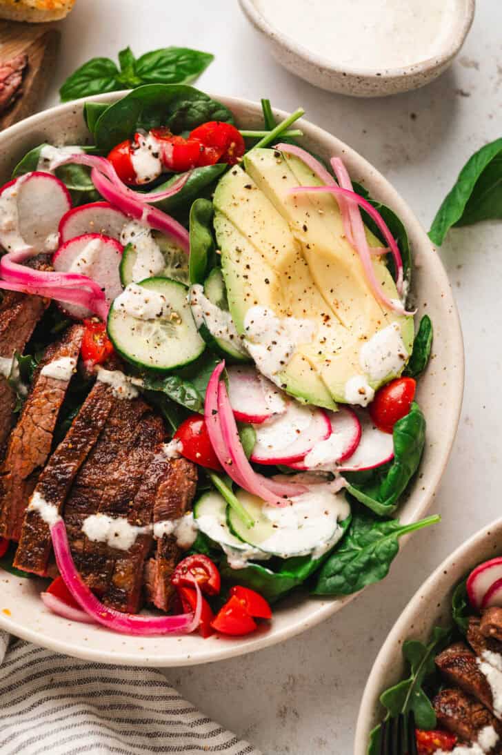 A bowl filled with steak salad with avocado, tomatoes, radishes, cucumber and pickled red onions.