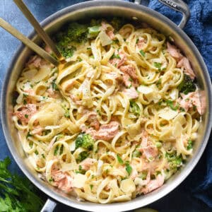 A large stainless steel skillet filled with salmon alfredo pasta and broccoli.