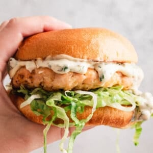 A hand holding a salmon burger in front of a light gray background.