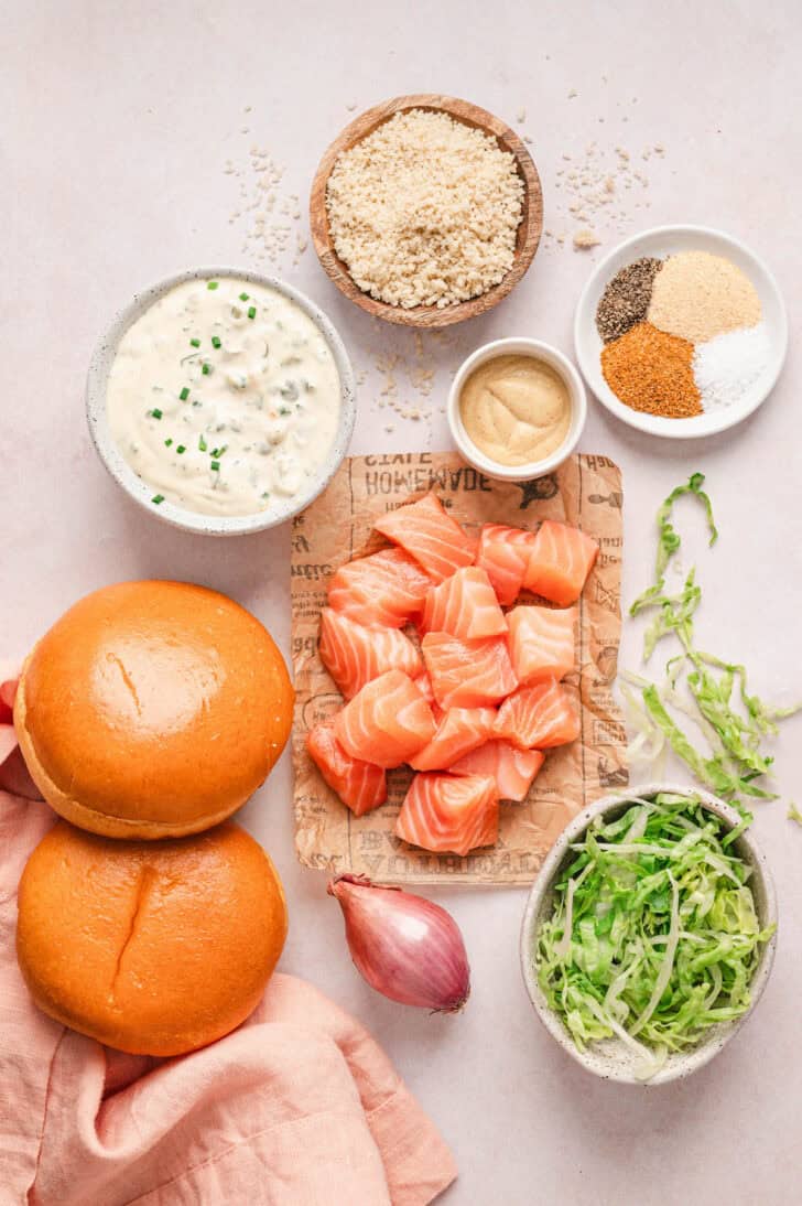 Ingredients on a light background, including cubed pink fish, tartar sauce, breadcrumbs, brioche buns, lettuce, mustard, shallot and spices.