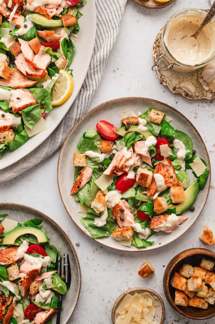 A salmon Caesar salad recipe in a dinner scene, with a large platter and two smaller plates of salad alongside extra dressing, cheese and croutons.