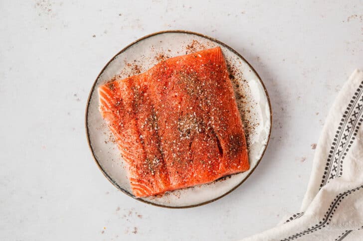 A piece of raw pink fish on a plate, sprinkled with spices.