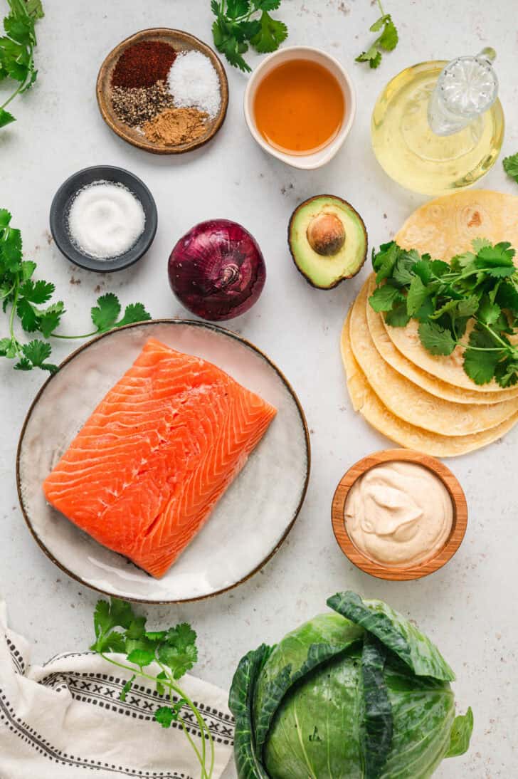 Ingredients on a white surface, including pink fish, tortillas, herbs, avocado, spices, onion, oil, cabbage and creamy sauce.