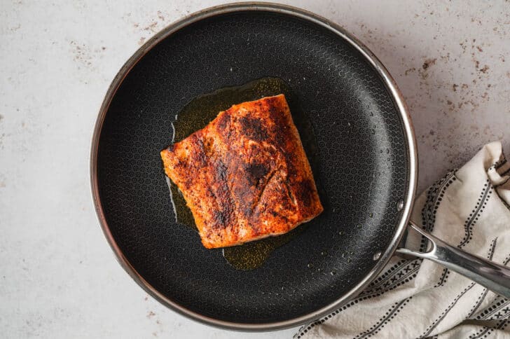 A spice rubbed piece of salmon cooking in a black skillet.