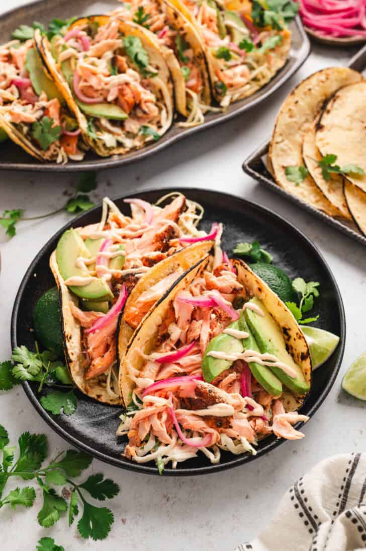 Two salmon tacos on a black plate in front of a platter of several more tacos.