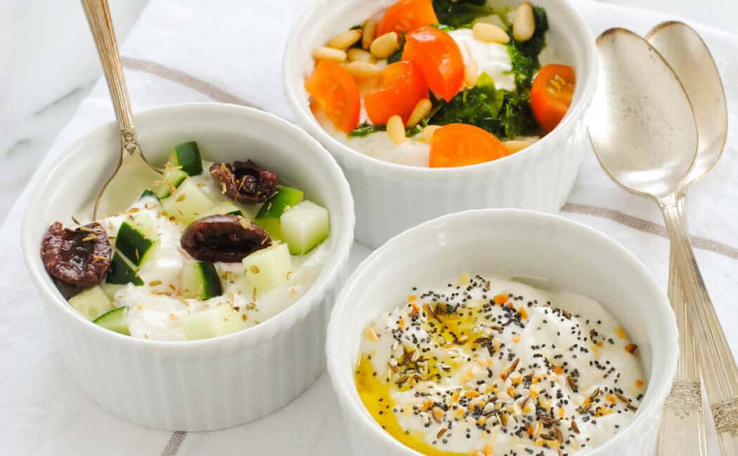 Three white ramekins of savory Greek yogurt recipes, including bowls topped with everything bagel seasoning, olives and cucumbers, and tomato, basil pesto and pine nuts.