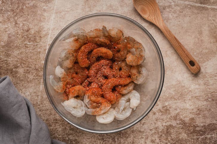 A glass bowl filled with raw shrimp topped with a red seasoning mix.