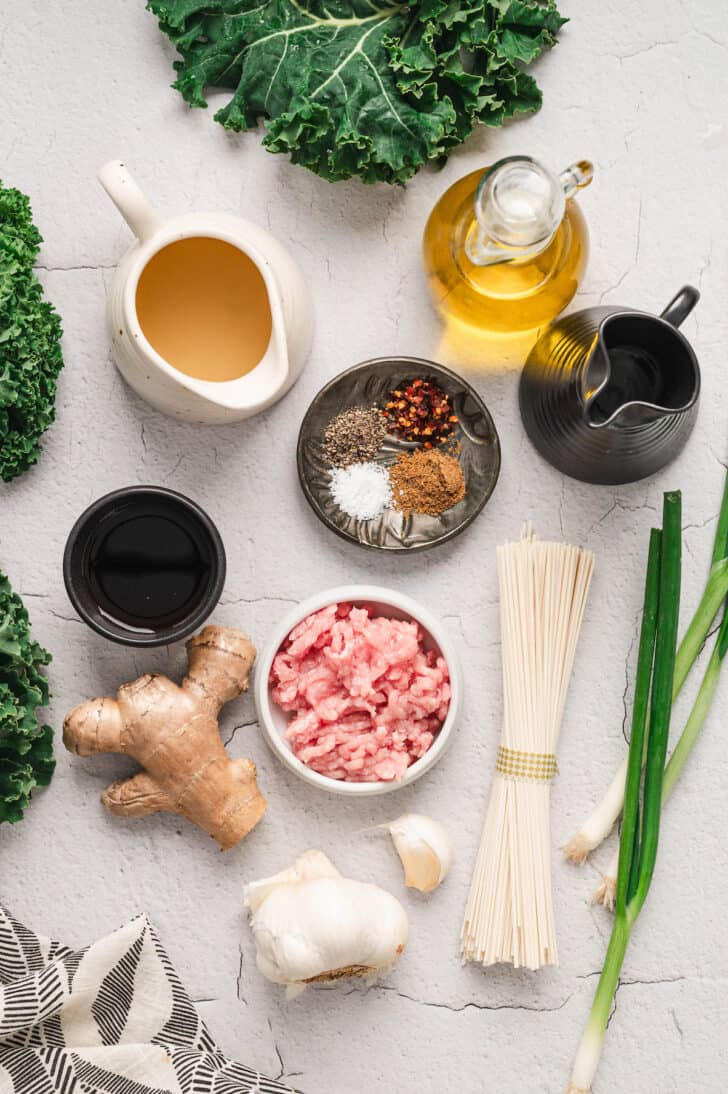 Ingredients on a white surface, including ground meat, broth, kale, green onions, udon, ginger, garlic, soy sauce and spices.