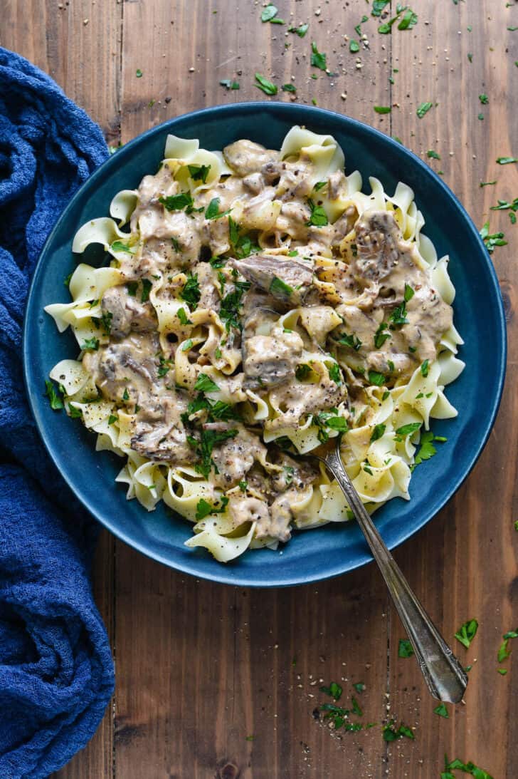 Creamy beef stew over egg noodles, served in a blue bowl with a spoon scooping a bite.