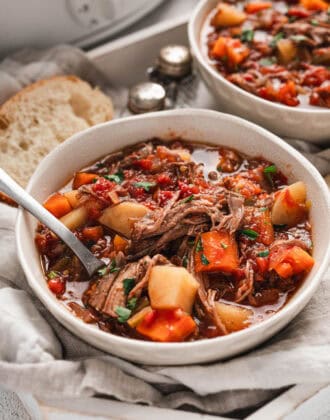 White bowls of crockpot vegetable beef soup in front of a slow cooker.