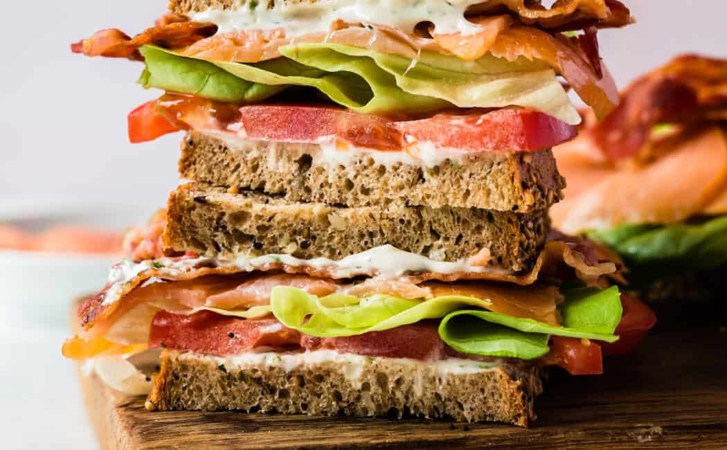 A stack of two smoked salmon BLT sandwich halves on a wooden cutting board in front of a light pink backdrop.