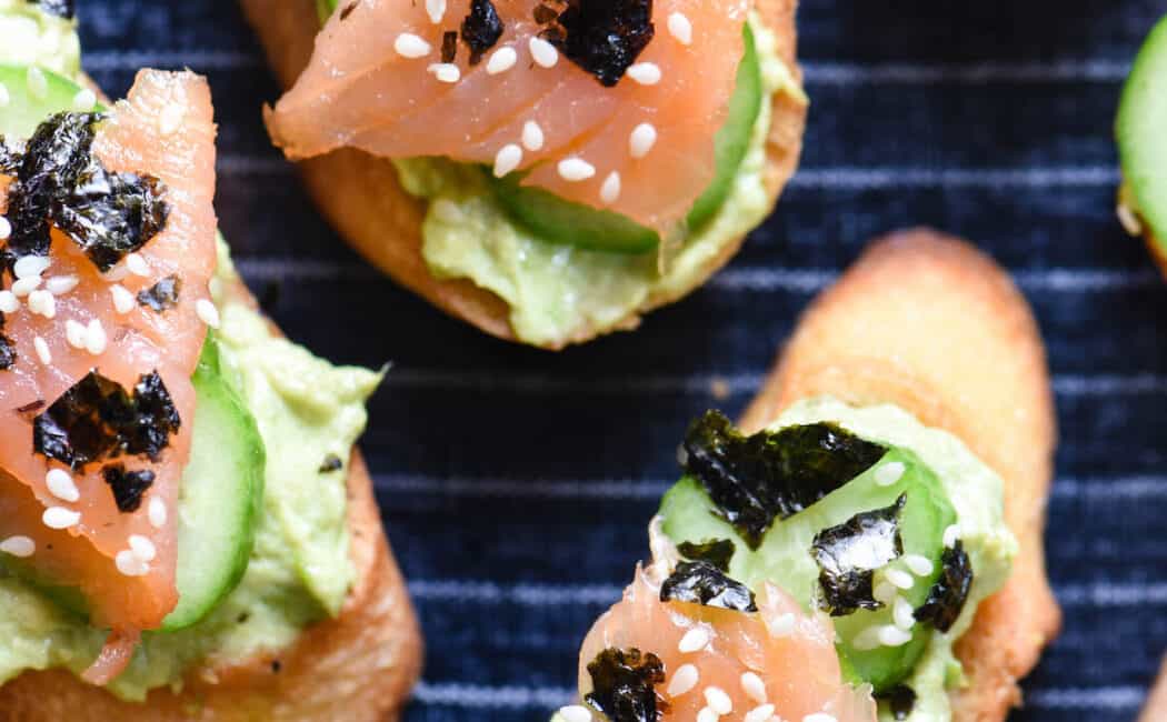 Toasted baguette topped with avocado spread, cucumber and smoked salmon, on top of blue and white striped serving tray.