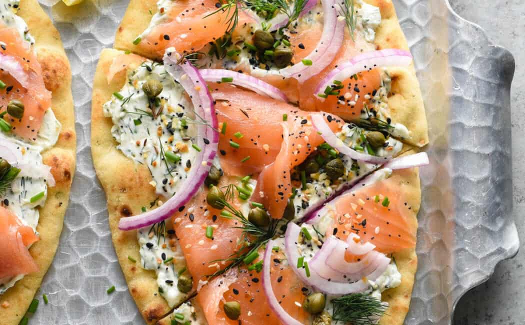 A smoked salmon pizza on a metal textured tray made with naan bread, cream cheese, herbs, everything bagel seasoning, lox, capers and red onion.