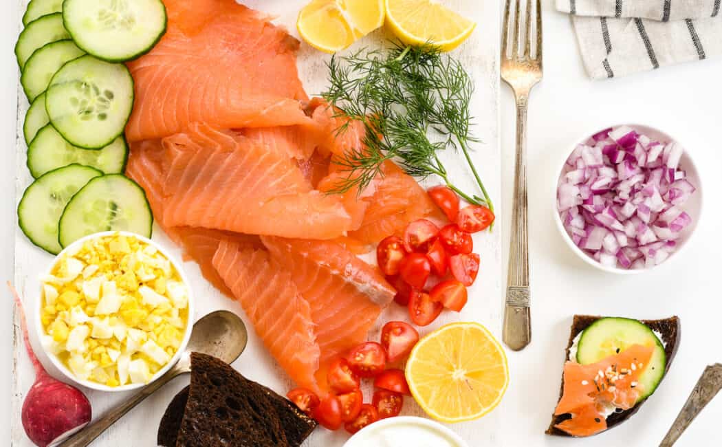 White table and cutting board topped with smoked salmon and garnishes like cucumber, capers, vegetables, cream cheese, bread and crackers.