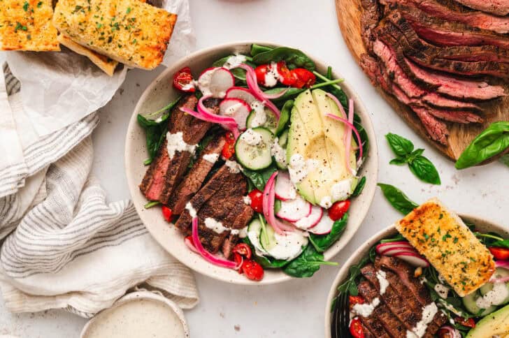 A dinner scene with bowls of steak and salad alongside sliced steak, pickled red onions and garlic bread.