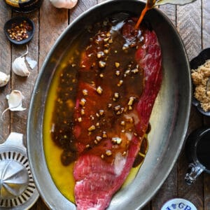 A piece of beef in a oval pan, with a hand pouring balsamic steak marinade with flecks of garlic in it over the meat.