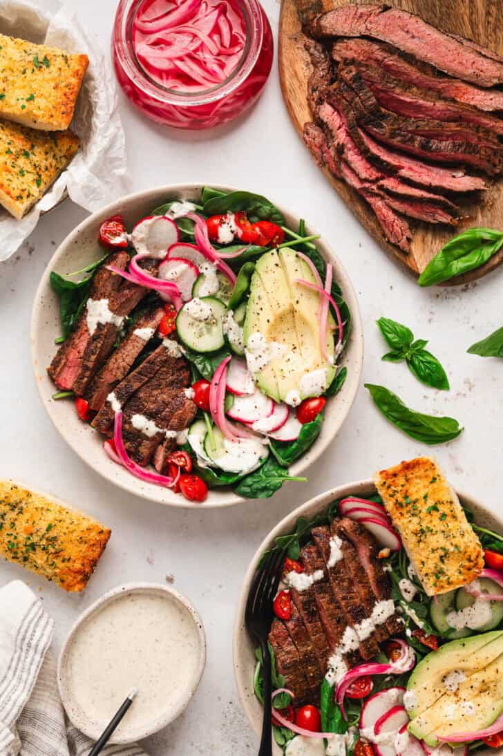 A dinner scene with bowls of a steak salad recipe alongside sliced steak, pickled red onions and garlic bread.