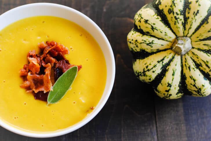 A small bowl of bright orange soup garnished with bacon and a sage leaf, on a dark table next to a whole dumpling squash.