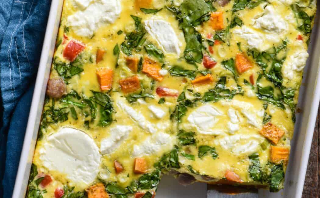 Sweet potato breakfast casserole made with eggs, sausage, spinach and goat cheese, in rectangular white baking dish, with a serving spatula lifting a piece out.