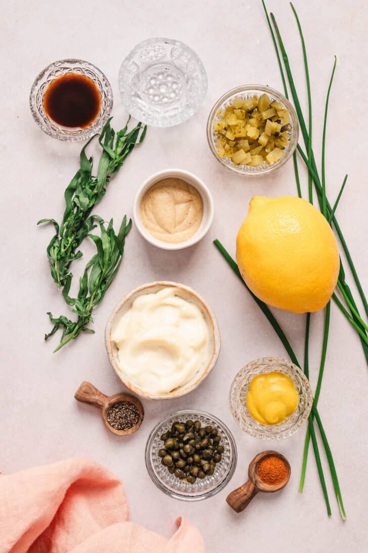 Ingredients on a light surface, including herbs, lemon, mayonnaise, mustard, chopped pickles, capers and spices.