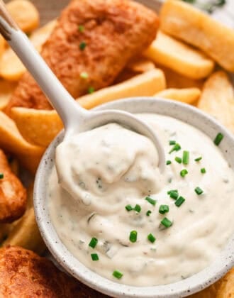 A spoon lifting a serving of a tartar sauce recipe from a white bowl, surrounded by fried fish and potatoes.