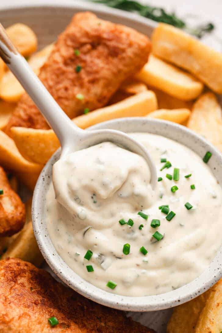 A spoon lifting a serving of a tartar sauce recipe from a white bowl, surrounded by fried fish and potatoes.