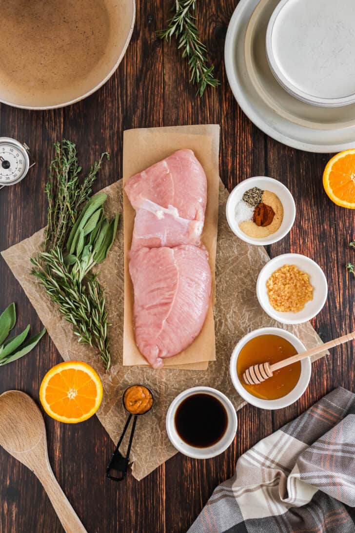 Ingredients on a wooden table, including raw poultry, spices, fresh herbs, honey, soy sauce, garlic, oranges and mustard.