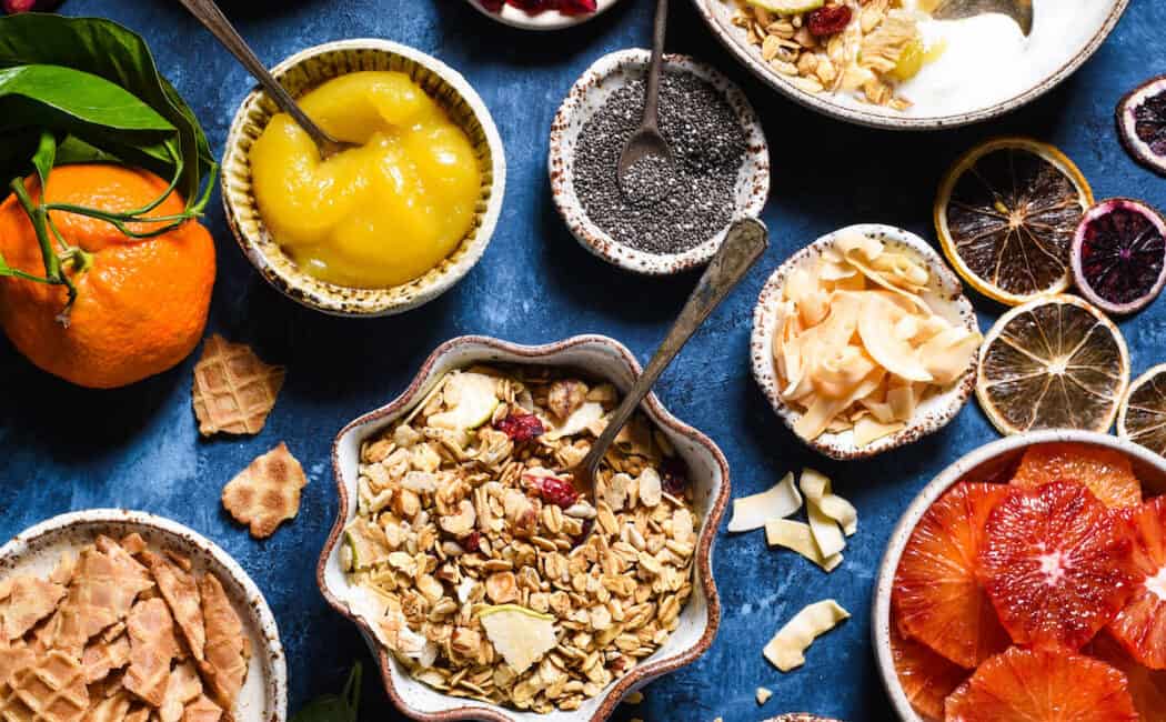 A yogurt bar set up on a blue background, with bowls of toppings including granola, waffle cone chunks, lemon curd, fruit, coconut, oranges and chocolate chunks.